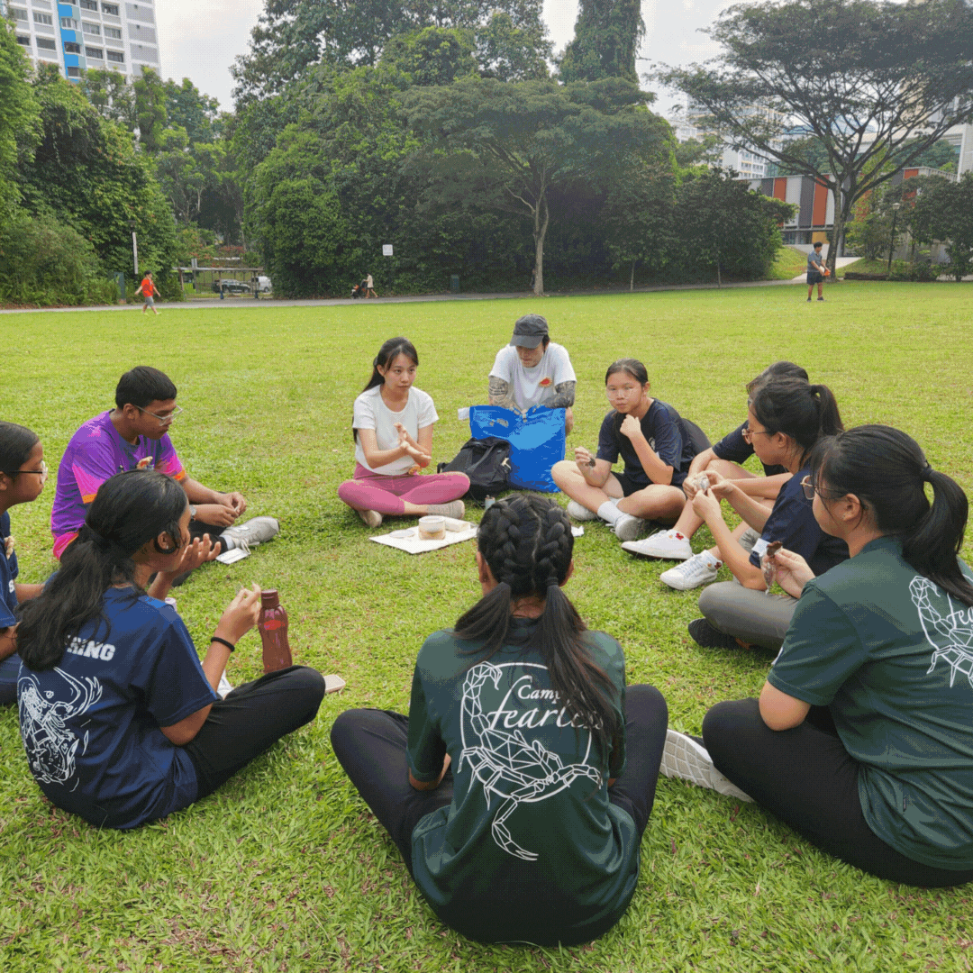 A group of about ten young people sit in a circle on grass in a park, engaged in an outdoor activity.